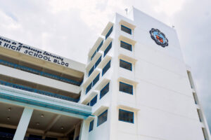 White high school building with blue-framed windows and a circular crest on the upper wall, viewed from an angled perspective.