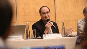 Man in a suit speaks into a handheld microphone at a panel table during a conference.