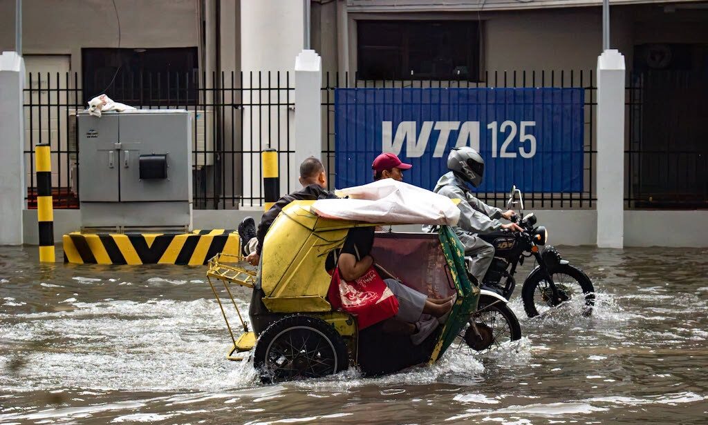 Motorcycle rider pulls a yellow improvised boat cart through waist-deep floodwater, with passengers inside the cart as water splashes around a gated street backdrop.