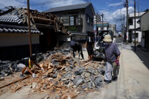 People clean up rubble from a collapsed roof on a residential street after a disaster, dragging tiles and wood amid shattered debris.