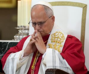 Pope in red robes sits in a gilded chair with hands folded in prayer, wearing a white veil and glasses.