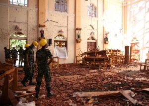 Soldiers in camouflage and yellow helmets survey a damaged church interior with broken pews and brick debris on the floor.