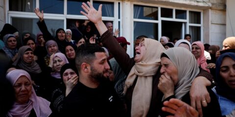 Crowd of grieving people, many women wearing headscarves, gathering outside a building with raised hands and somber expressions.