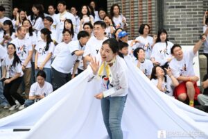 Woman in a white T-shirt waves from the edge of a large white banner while a cheering crowd in matching shirts stands behind her.