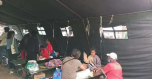 Inside a dark canvas aid tent, several people sit or lie on benches and cots while volunteers provide care at a makeshift clinic.